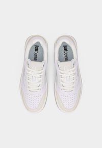 Ζευγάρι λευκά αθλητικά παπούτσια με κορδόνια, με διαμπερές τμήμα στα δάχτυλα και μπεζ λεπτομέρειες, φωτογραφημένα από πάνω, με το λογότυπο "Just Cavalli" ορατό στις εσωτερικές σόλες.