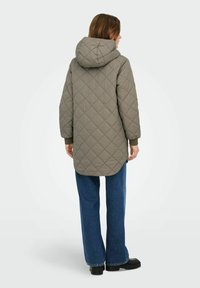 Prešit parka jakna olivno zelene barve z rancljem, z dolgimi rokavi in daljšo zadnjo stranjo, kombinirana z ohlapnimi modrimi jeans hlačami in črno obutvijo.