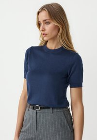 Marineblaues, gestricktes Kurzarmoberteil mit Rundhalsausschnitt, glatter Textur und taillierter Passform, kombiniert mit grauen Nadelstreifenhosen und einem schwarzen Gürtel.