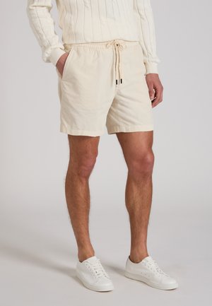 Homme portant un short crème à cordon, des baskets blanches et un pull en maille torsadée crème, debout avec une main dans la poche.