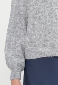 Pulover gri din tricot cu un model texturat, tip cablu, și mâneci bufante. Tivul de jos este  ribat. Purtați peste un articol de îmbrăcăminte de culoare albastră închisă.
