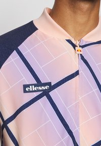 Trekkjakke med et rosa og marineblått geometrisk mønster, med en logo-patch og glidelås. Mykt stoff med teksturert krage.
