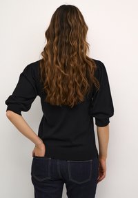 Pull en tricot noir avec des manches bouffantes, cintré à la taille, associé à un jean en denim foncé. Cheveux ondulés tombant au-delà des épaules.