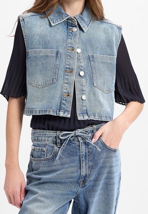 Frau trägt eine hellblaue ärmellose Jeansweste über einem schwarzen plissierten Kurzarm-Top, kombiniert mit hoch taillierten Jeans mit gebundenem Gürtel.