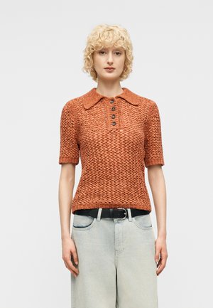 Άτομο με κοντά σγουρά ξανθά μαλλιά, που φοράει ένα υφασμάτινο, σκουροπορτοκαλί πλεκτό κορμάκι με κουμπιά και ανοιχτόχρωμο φαρδύ τζιν.