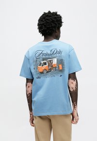 Gaiši zils kokvilnas T-krekls ar oranžas pārtikas kravas automašīnas grafiku un tekstu "Fresh Daily" mugurpusē, īsām piedurknēm un apaļu kakla izgriezumu.