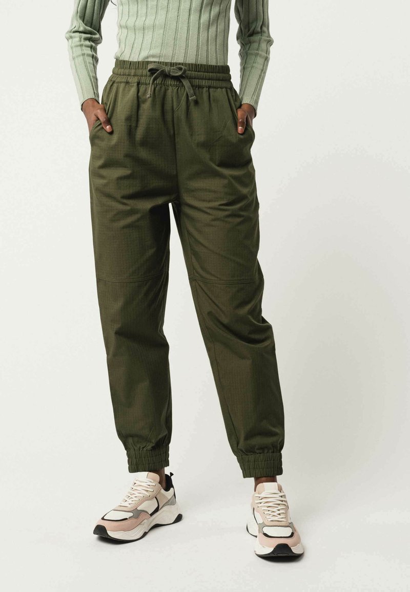 Olijfgroene, lichte broek met elastische tailleband en boorden. Heeft twee zijzakken en een gladde textuur. Draagt met trendy sneakers.