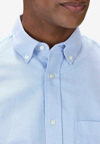 Vaaleansininen nappikauluspaita, jossa on terävä kaulus, valkoiset napit ja rintatasku. Valmistettu teksturoidusta kankaasta.