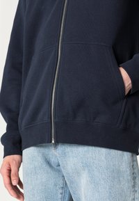 Felpa con zip blu navy con finitura testurizzata, tasche a canguro, polsini e vita a costine, abbinata a jeans azzurri chiari.