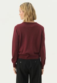 Burgunderfarbener Pullover mit Rundhalsausschnitt und langen Ärmeln, aus weichem Stoff. Getragen mit schwarzen Hosen, die über zwei Gesäßtaschen verfügen.