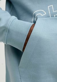 Lichtblauwe sweatshirt met een geribbeld manchet. De stof is glad met een kangoeroezak; de zichtbare hand heeft een donkerder huidskleur.