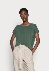 Green short-sleeve t-shirt in a soft material with a relaxed fit, round neckline, and rolled sleeves, paired with light beige trousers.