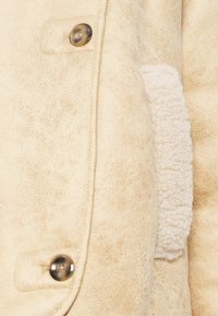 Veste en simili cuir beige avec une finition texturée, dotée d'une doublure en crème duveteuse et de deux boutons ronds en métal à l'avant.