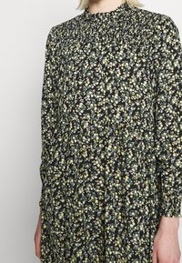 Svart blommig klänning med en rynkad halsringning, långa ärmar och en flödande silhuett med små gula blommor och gröna blad.