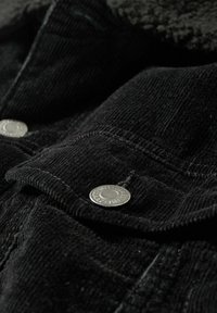 Sort cordurojakket med sølvmetal knapper, tekstureret stof og en blød, pelsagtig krave. Fokus på syninger og knapplacering.