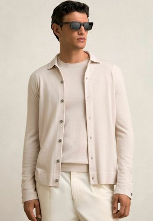 Cardigan leggero beige chiaro con bottoni, indossato sopra una camicia crema abbinata. Modello con occhiali da sole neri, in posa su uno sfondo neutro.