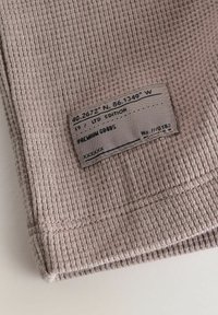 Tkanina s textúrou vaflí v tlmenej marhuľovej farbe s našitou etiketou, ktorá uvádza súradnice, limitovanú edíciu a podrobnosti o značke.