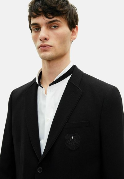 Schwarzer Blazer mit strukturiertem Finish, mit einem schwarzen Emblem auf der Tasche und einem weißen Hemd mit kontrastierendem schwarzen Kragen.
