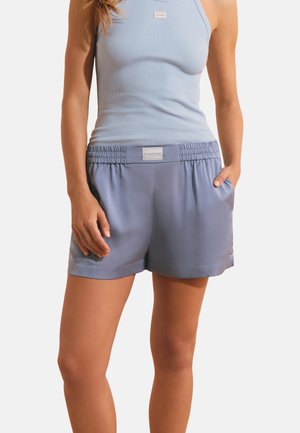 Frau trägt hellblaues geripptes Tanktop und passende Shorts mit elastischem Bund und dem Aufdruck "Week End Club", steht mit den Händen in den Taschen.