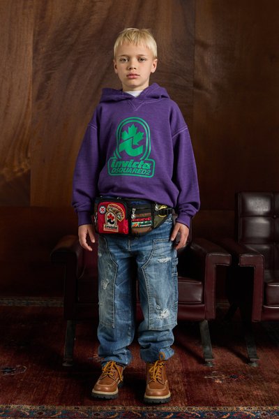 Felpa viola con logo verde, jeans blu strappati, stivali marroni stringati e una cintura colorata con vari toppe.