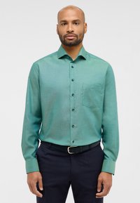Camicia verde con bottoni, taschino a sinistra, maniche lunghe e bottoni scuri, realizzata in un tessuto liscio, indossata con pantaloni scuri.