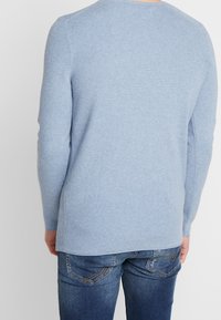 Ljusblå stickad tröja med långa ärmar, rund halsringning och texturerat mönster. Bärs över mörk denimjeans, vilket ger en avslappnad passform.