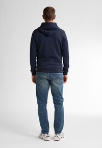 Navy blue hoodie with a hood, ribbed cuffs, and hem, paired with light blue distressed jeans, and white athletic shoes.