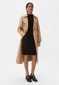 Trench-coat beige, design double boutonnage, avec grands revers et ceinture. Robe noire ajustée en dessous, associée à des mocassins en cuir verni.