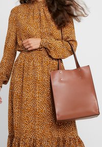 Brązowa torba typu tote o gładkiej fakturze, z dwoma uchwytami u góry i subtelnymi metalowymi akcentami. Noszona z brązową sukienką we wzory, z marszczonym dekoltem.