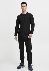 Black textured long-sleeve shirt with ribbed cuffs, paired with black pants and navy athletic shoes, rolled cuffs visible, standing pose.