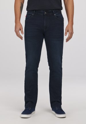 Mężczyzna ubrany w ciemnoniebieskie jeansy typu slim-fit i granatowe trampki, stojący na tle o neutralnym kolorze.