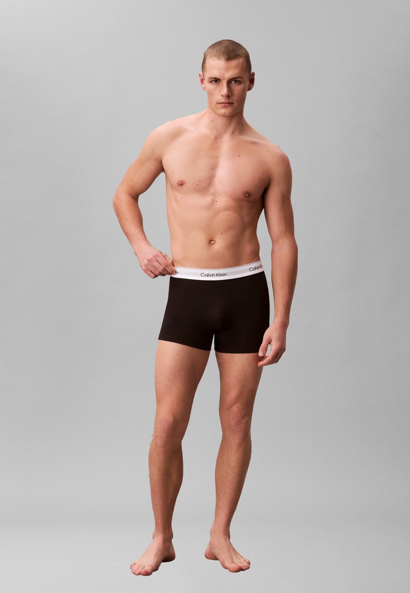 Junger, oberkörperfreier Mann in schwarzen Calvin Klein Boxershorts steht vor grauem Hintergrund, hält mit der linken Hand den Hosenbund.
