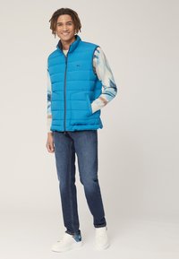 Gilet blu imbottito, con chiusura a cerniera, indossato da un modello che porta una camicia a maniche lunghe con stampa, jeans e sneakers bianche con dettagli colorati.