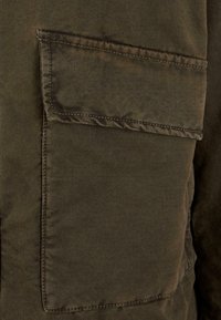 Olivgrüne Jacke mit einer großen aufgesetzten Tasche. Hergestellt aus strapazierfähigem, strukturiertem Stoff, mit kontrastierenden Nähten entlang der Kanten.