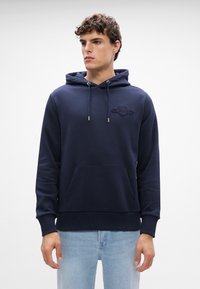 Námornícky modrá mikina s kapucňou vyrobená z mäkkého materiálu, má predné vrecko, nastaviteľné šnúrky a vyšívané logo na hrudi.