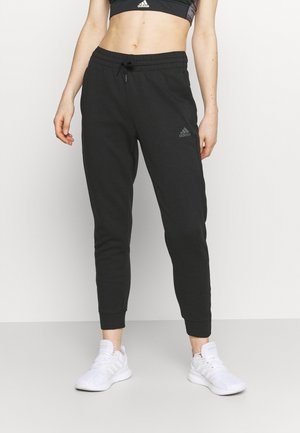 Persoon die zwarte Adidas joggers, witte Adidas sneakers en een zwarte Adidas sportbeha draagt, staand tegen een effen lichte achtergrond.