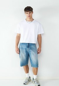 Biała koszulka z krótkim rękawem zestawiona z jasnoniebieskimi dżinsowymi szortami. Model nosi szare trampki z białymi skarpetami. Minimalistyczne tło.