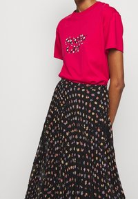 Fuchsia T-shirt med "Seedy Girl" grafik, parret med en sort plisseret nederdel med et multifarvet blomstermønster.