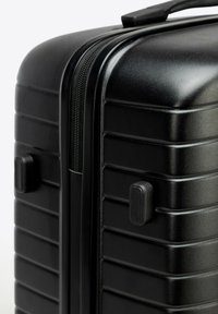 Esquina de maleta rígida negra con cremallera y asa, presentando ranuras horizontales y una traba en el lateral.