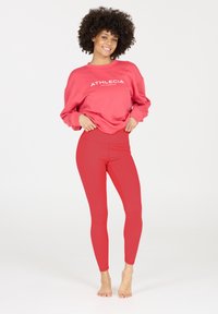 Rode sweatshirt met witte tekst "ATHLECIA" en rode high-waisted leggings, gladde textuur, relaxte pasvorm, gecombineerd met blote voeten op een neutrale achtergrond.