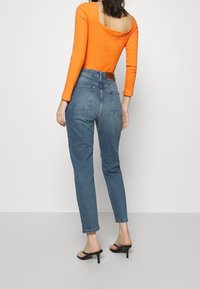 Frau in hochgeschnittenen blauen Jeans, orangefarbenem Langarmtop mit offener Rückseite und schwarzen Absatzsandalen, die vor einem hellen Hintergrund steht.