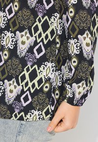 Blusa a maniche lunghe in tessuto nero con un motivo astratto multicolore, che include quadrati, spirali e disegni paisley. Texture morbida.