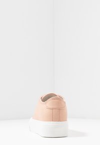 Jasnoróżowy płócienny trampki pokazane od tyłu z grubą białą gumową podeszwą na białej powierzchni i tle.