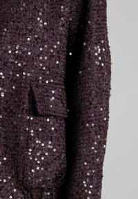 Giacca in tessuto strutturato viola scuro decorata con piccole paillettes lucide, dotata di una tasca a patta e maniche lunghe su uno sfondo semplice.