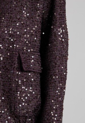 Chaqueta de tela texturizada de color púrpura oscuro adornada con pequeños brillantes, con un bolsillo tipo solapa y mangas largas, sobre un fondo liso.