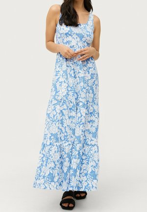 Robe maxi en tissu bleu clair avec des motifs floraux blancs, épaules dénudées et design à jupe à volants. Portée avec des sandales noires.
