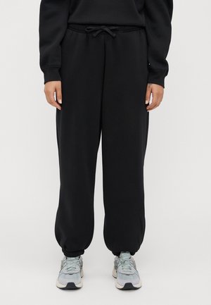 Person med svarta joggers med elastiskt midjeband och avklippta anklar, i ljusgrå sneakers, stående mot en plain bakgrund.