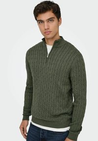 Pull en maille torsadée vert avec un col zippé à mi-hauteur. Cuffs et ourlet côtelés, tissu texturé, porté par-dessus une chemise blanche.