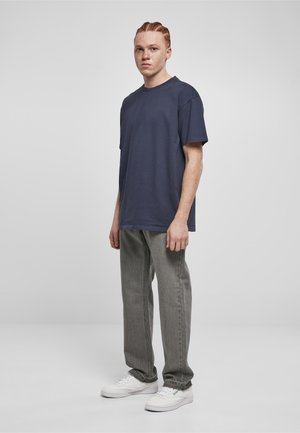 Jeune homme aux cheveux tressés portant un t-shirt bleu marine, un jean gris et des baskets blanches, debout devant un fond clair uni.