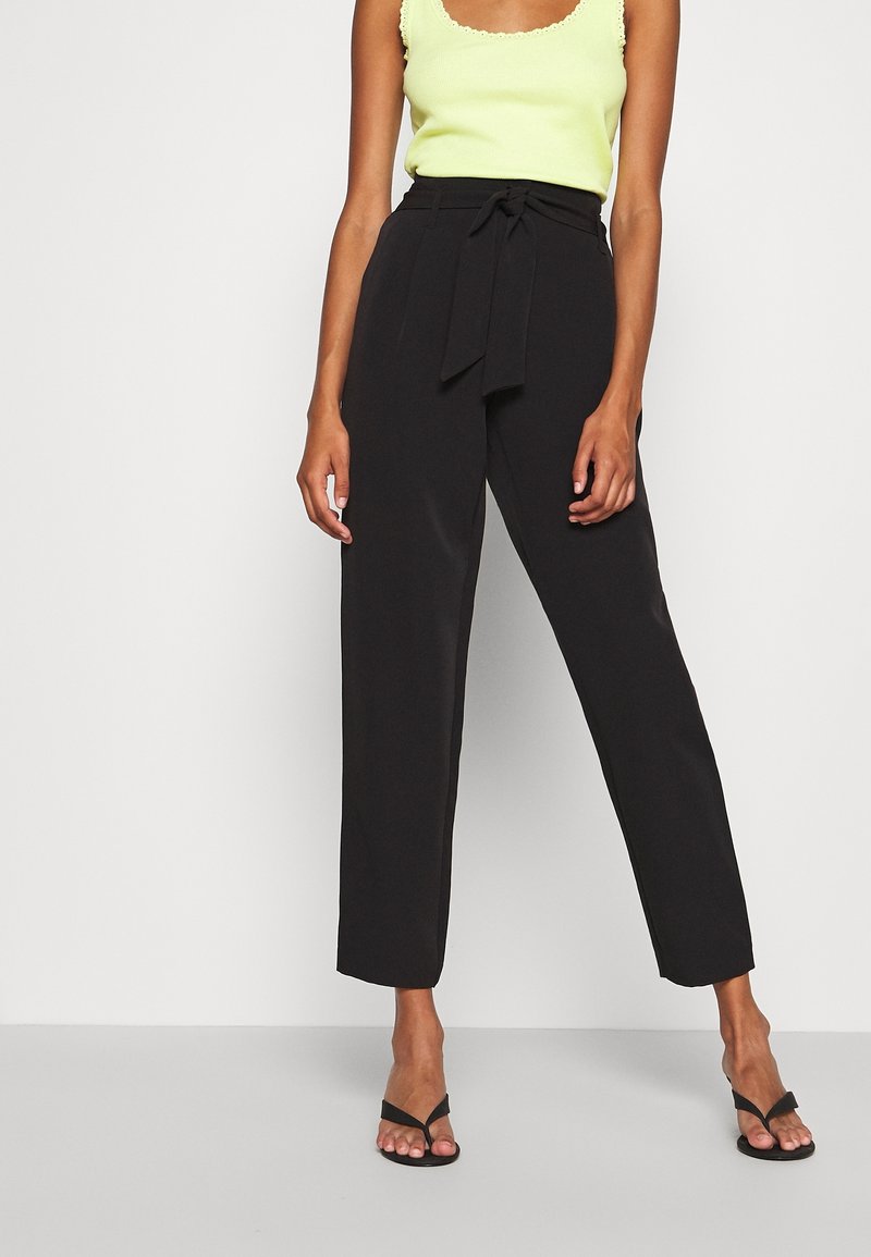 Femme portant un pantalon noir taille haute style paperbag avec une ceinture nouée, associé à un haut sans manches vert clair et des chaussures à talons ouvertes noires.